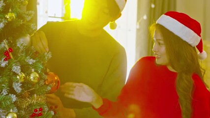Asian couple wearing santa hat decorates christmas tree with bauble ornament and warm sunlight creating cozy festive mood young woman red sweater and man hanging red ornament christmas tree smiling - Powered by Adobe
