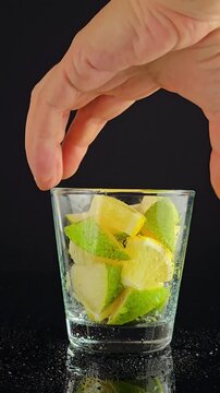 Hand taking a glass with fruit pieces of lemon and pear on a dark background in vertical video. person grabbing a glass filled with cut fruit for smoothie bars, fruit salad recipe, healthy food