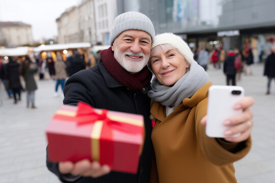 photo of a happy senior couple taking a selfie while holding christmas gifts at the city square during winter, with many people around and a shopping center in the background. capt