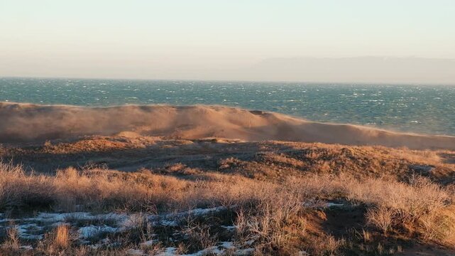 A windy shore, a drift of sand rises above the dunes against the background of a stormy sea in the rays of the setting or rising winter sun.