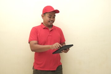 Cheerful Southeast Asian man in red shirt and cap using tablet, isolated with copy space, ideal for...