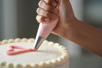 Person's hand decorating a white cake with pink buttercream heart frosting, a sweet and delicate baking process in action