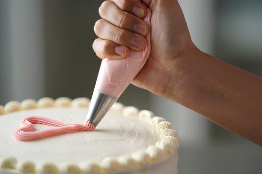 Person's hand decorating a white cake with pink buttercream heart frosting, a sweet and delicate baking process in action