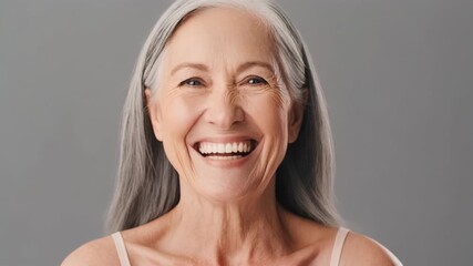 Radiant older woman with silver hair smiles joyfully against a gray backdrop, showcasing positive emotion and aging gracefully in studio.