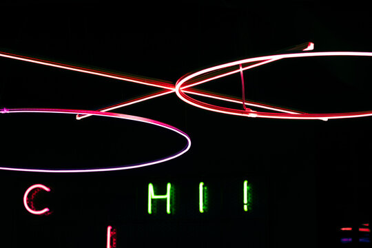 abstract composition featuring bright neon light rings and linear elements in pink, yellow, and white against a dark background. below the installation, green and red illuminated letters add extra dep