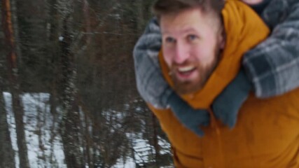 Cheerful Caucasian boy riding on father back along snowy forest path enjoying playful outdoor moment on cold winter day