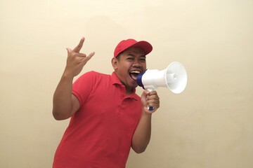 Energetic Southeast Asian man in red shirt and cap shouting into megaphone with rock gesture,...