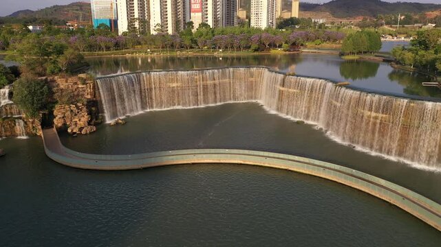 Kunming, China - 06 December 2025: Aerial View of a constructed waterfall feature cascading into a pool of water, framed by lush trees and distant high-rise buildings on a sunny day.