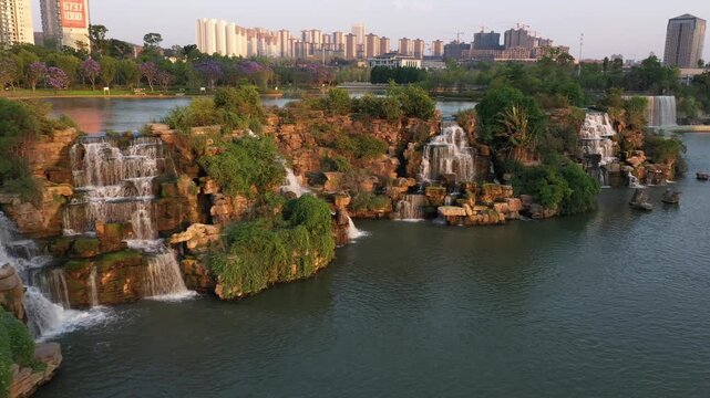 Kunming, China - 06 December 2025: Aerial View of tiered waterfalls cascade amidst lush greenery, set against a backdrop of modern city buildings under a soft, golden sky.