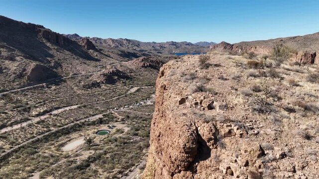 Aerial view of rugged mountains and valley, punctuated by sparse vegetation and a distant lake under a clear sky, Apache Junction, Arizona, United States.