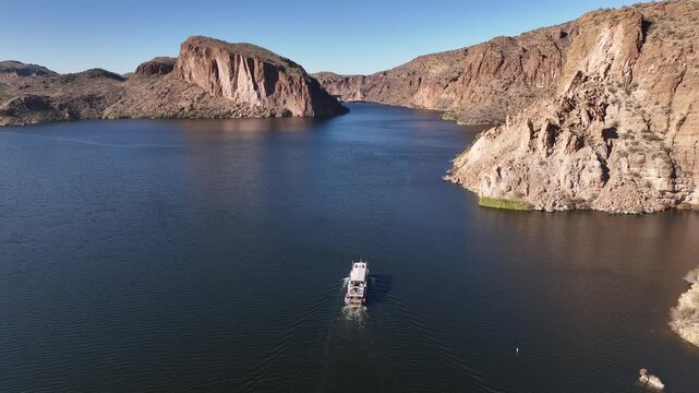 Aerial view of a boat cutting through the tranquil, dark blue waters amidst rugged mountains under a bright blue sky, Apache Junction, Arizona, United States.