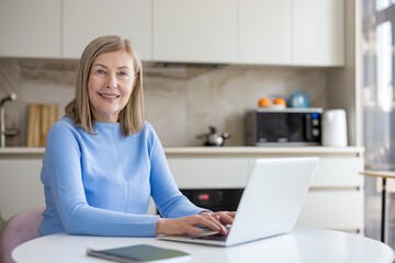 Senior woman smiling while typing on laptop at kitchen table, working remotely and learning online-confident, connected, and enjoying modern digital, freelance lifestyle at home
