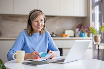 Senior woman is participating in an online meeting or e-learning session from her home kitchen, actively engaging with technology while wearing a headset and taking notes in a planner