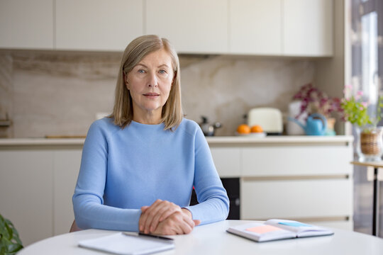 Mature businesswoman sitting at a table in her kitchen, engaging in a corporate video conference, representing remote work and freelance opportunities for older professionals
