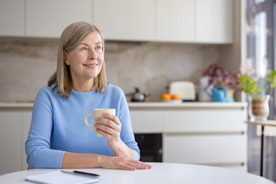 Senior woman relaxing at the kitchen table, holding a coffee mug, looking away with a thoughtful expression, enjoying a quiet moment at home during the morning - Powered by Adobe