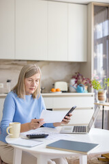 Senior woman at home checking documents and using a laptop and smartphone to calculate expenses,...