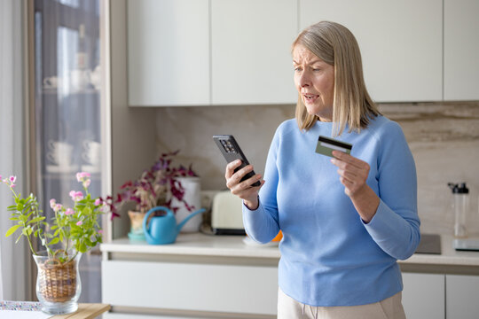 Senior woman looking confused while holding a smartphone and credit card, experiencing difficulty with an online transaction or payment in a modern kitchen setting - Powered by Adobe