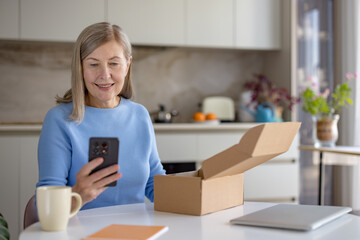 Senior woman smiling, holding smartphone, and looking at a cardboard delivery box, excited about...