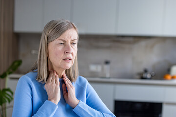 Senior woman suffering from a sore throat or thyroid discomfort, touching her neck and showing a pained facial expression due to illness and swelling while at home