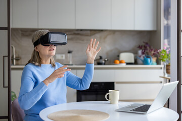 Senior woman wearing a vr headset and interacting with virtual space, sitting at a kitchen table with a laptop, embracing modern technology and the metaverse