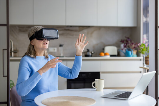 Senior woman wearing a vr headset and interacting with virtual space, sitting at a kitchen table with a laptop, embracing modern technology and the metaverse