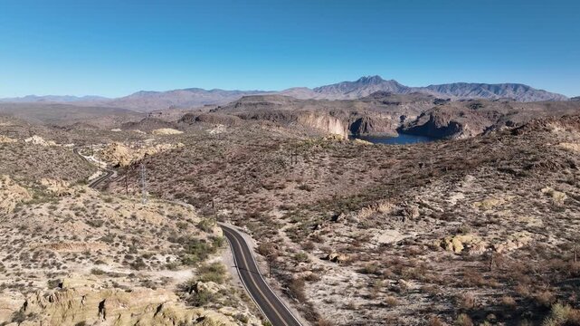 Aerial view of a winding desert road and a serene lake nestled amid arid mountains, creating a striking contrast of colors and textures, Apache Junction, Arizona, United States.