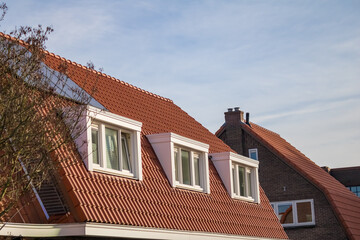 Dormer windows on a traditional brick building with tiled roofs, featuring contrasting red and neutral window frames reflecting autumn trees. High quality photo