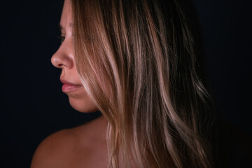 Fototapeta premium Side close-up of a young woman with healthy shiny blonde hair, soft lips and natural skin texture, posing on a dark background in gentle studio light