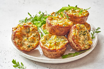 Savory zucchini corn egg muffins are freshly baked and beautifully arranged on a white plate. The muffins are surrounded by vibrant greens, adding a touch of freshness to the scene.