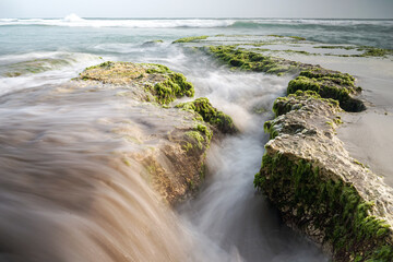 Ethereal coastal scenery featuring ocean water cascading over moss-covered rocks under a soft, overcast sky offering a serene and timeless view of natural beauty