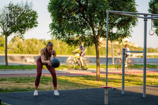 Concentrated sportswoman practicing functional training with medicine ball in a city park during a sunny day, improving balance, coordination, and core strength