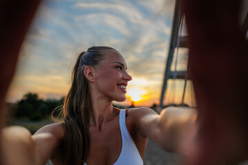 Young, smiling sportswoman capturing a selfie at sunset on a bridge, savoring the golden hour after an invigorating workout, radiating joy and positivity in her fitness journey