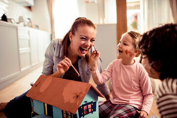 Adult mother and child daughter laughing while painting cardboard house at home