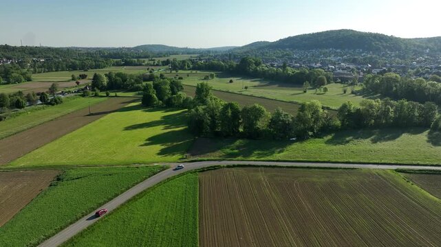 Aerial view of lush green and brown fields interspersed with trees, contrasting with the distant settlement nestled against the hills, Zillisheim, Grand Est, France.