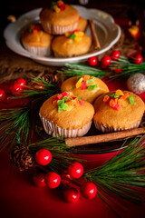 A warm and festive still life of Christmas-themed cupcakes topped with colorful candied fruit. The cupcakes are arranged with holiday decorations including pine branches, red berries, and rustic.