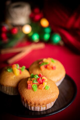 A warm and festive still life of Christmas-themed cupcakes topped with colorful candied fruit. The cupcakes are arranged with holiday decorations including pine branches, red berries, and rustic.