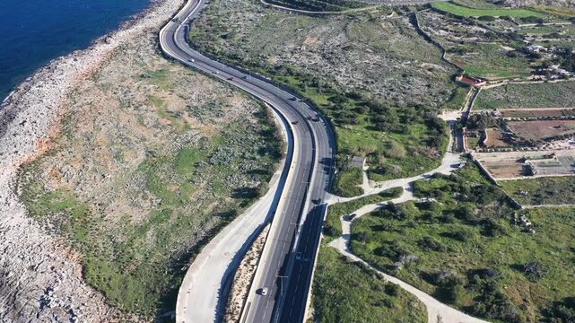 Aerial view of a winding road cutting through a rocky coastline with the deep blue sea contrasting with the arid landscape, St. Paul's Bay, Malta.