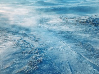 An aerial view reveals a vast, frozen landscape with thick snow cover showing intricate wind-blown patterns, undulating snowy foothills, and a misty, cold atmosphere under an overcast winter sky.