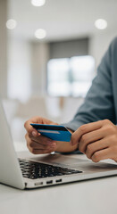 Fototapeta premium Close-up of a blue credit card held by a person near a laptop, representing online purchase, payment or e-commerce transaction