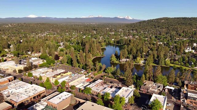 Aerial view of Mirror Pond's dark waters contrasting with the lush greenery and buildings, shadowed by distant, snow-capped mountains, Bend, Oregon, United States.