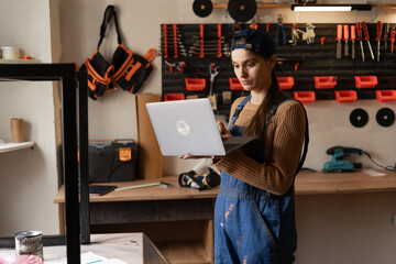 Woman carpenter in blue overalls typing laptop in workshop. worker artist using app online, Female craftsman