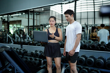 Asian man and woman exercise training in fitness gym. Young female trainer using laptop for schedule exercise training to fitness male. Workout training in fitness gym.