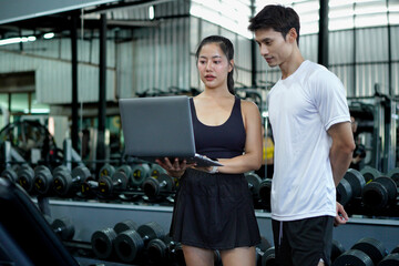 Asian man and woman exercise training in fitness gym. Young female trainer using laptop for schedule exercise training to fitness male. Workout training in fitness gym.