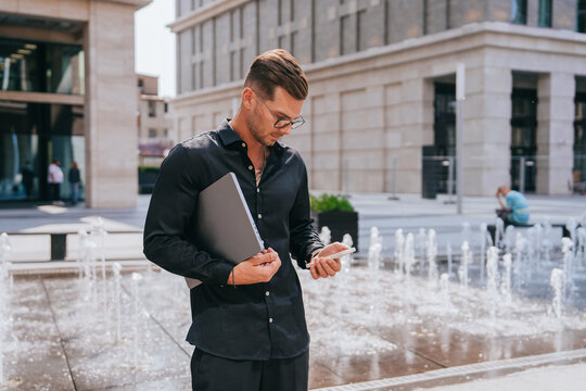 A man stands by a fountain checking his phone, holding a laptop, exuding a blend of professionalism and modern connectivity.