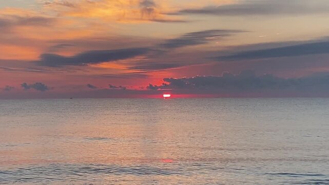 A tranquil scene at the beach with the sun rising above a calm ocean with small waves in the foreground and purple clouds in the sky for a beautiful coastal seascape