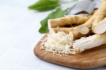 Grated horseradish, grater, leaves and fresh roots on white tiled table, closeup. Space for text