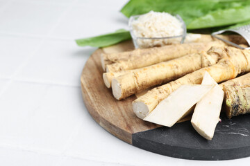 Whole and grated horseradish roots and green leaves on white tiled table, closeup. Space for text