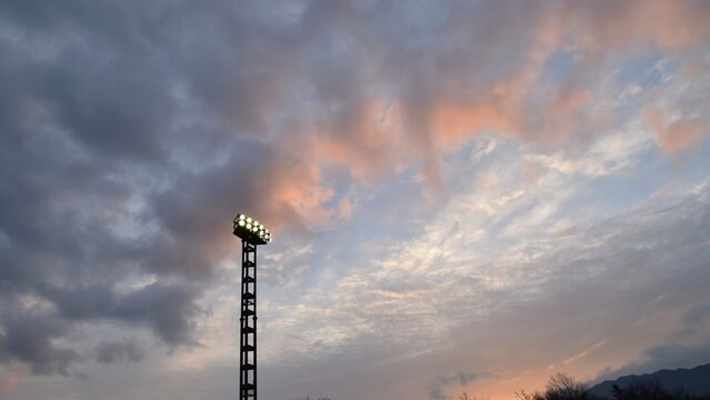 Dramatic sunset sky with pink and orange clouds illuminated above a tall stadium floodlight tower, capturing the atmosphere of an evening sports event about to begin.