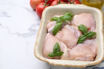 Raw chicken thighs with basil in baking dish on white marble table, closeup. Space for text