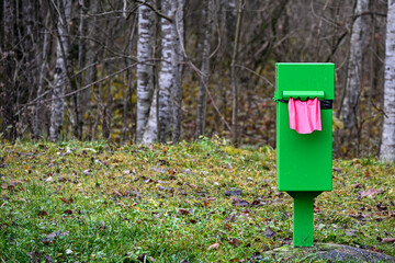 Bright green dog-waste bag dispenser standing in a grassy forest area, with a pink bag visible, creating a vivid contrast in a natural outdoor setting